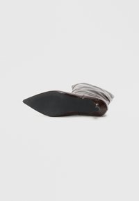 Bota de cuero marrón oscuro tumbada de lado, mostrando la suela puntiaguda negra con un agarre texturizado contra un fondo blanco liso.