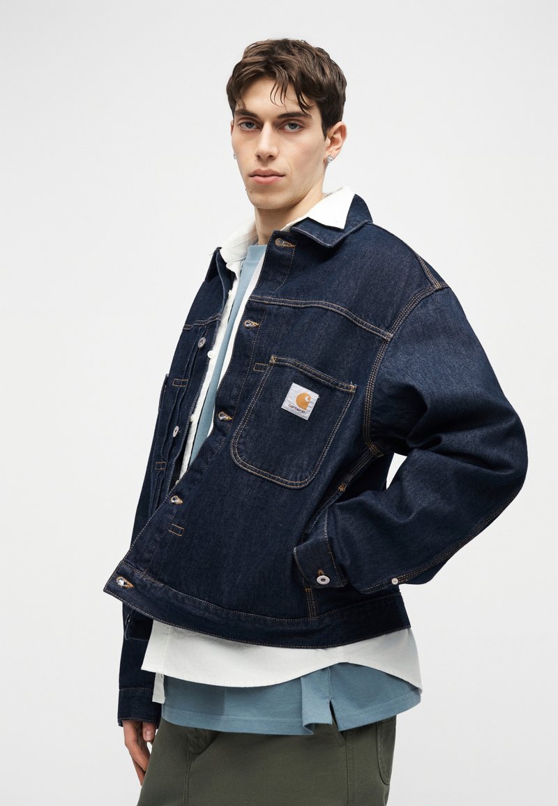 Junger Mann trägt eine dunkelblaue Jeansjacke mit weißem Kragen über mehreren blauen und weißen Hemden und steht vor einem schlichten weißen Hintergrund.