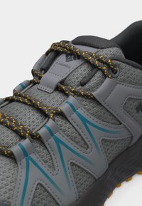 Pilko „Columbia“ žygių batelio su mėlynomis detalėmis ir juodomis raištelėmis geltonais taškeliais aprašymas, išsiskiriantis tekstūruotu audinio viršumi ir tvirtu padu.