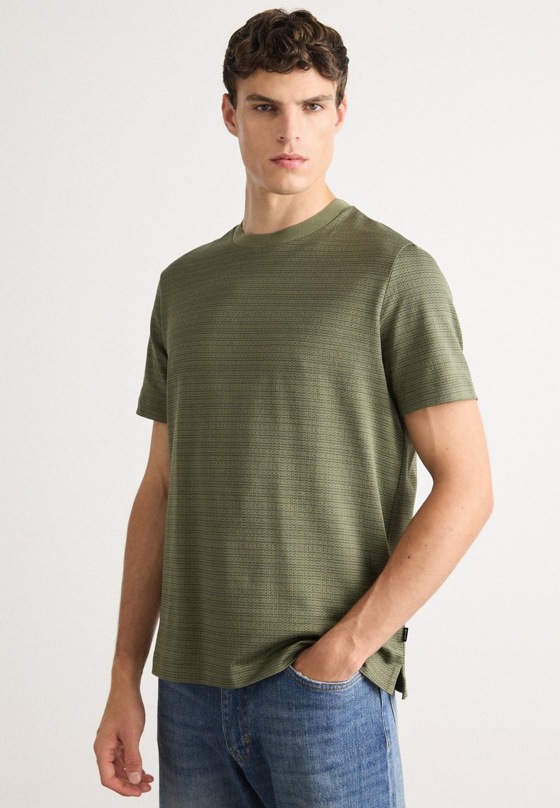 Mladenič oblečen v olivno zeleno teksturirano majico s kratkimi rokavi in modre kavbojke, stoji z roko v žepu pred belo ozadje.