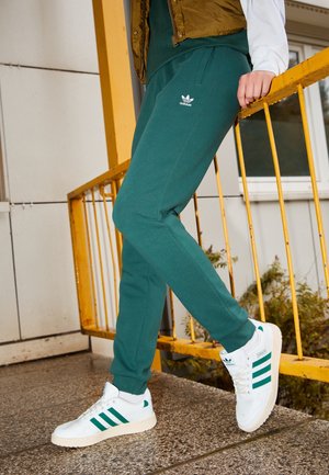 Osoba nosząca zielone dresy Adidas i białe sneakersy Adidas z zielonymi paskami, opierająca się o żółtą balustradę na zewnątrz.