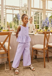 orgonalila kétrészes öltözék ujjatlan, gombos peplumfelsővel és bőszárú nadrággal, világos pántos szandállal párosítva.