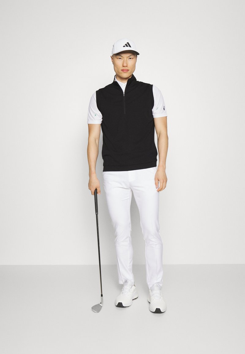 Zwarte ritsvest over een wit T-shirt met korte mouwen, gecombineerd met witte broek en witte sportschoenen. Houdt een golfclub vast.