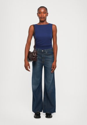 Navy ärmelloses Top kombiniert mit weit geschnittenen, dunklen Jeans. Die Accessoires bestehen aus einer kleinen, geflochtenen, dunkelbraunen Handtasche und schwarzen Schuhen.