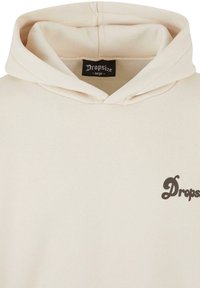Sweat à capuche beige avec une poche avant et un logo noir "Dropsize" sur la poitrine, ainsi qu'une étiquette de taille à l'intérieur de l'encolure, indiquant une taille large.