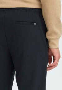 Mustat, teksturoidut housut takataskulla, jossa on pieni valkoinen logo. Materiaali näyttää pehmeältä ja istuvuus on rento. Käsi beige-syysvaatteessa.