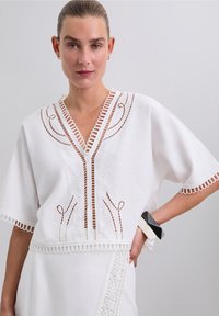 Haut en lin blanc avec un décolleté en V, présentant des motifs découpés intriqués et des manches à bord en dentelle. Accentué par un bracelet géométrique audacieux au poignet.