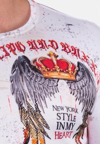 Weißes, eng anliegendes Hemd mit schwarzen Flügeln, einem roten Kronen-Graphic und dem Text "NEW YORK STYLE IN MY HEART" in fetter, roter Schrift. Mit Strasssteinen akzentuiert.
