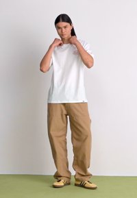 Un tricou alb din bumbac cu un logo mic, combinat cu pantaloni cargo khaki și adidași galbeni cu dungi negre, pe un fundal simplu.