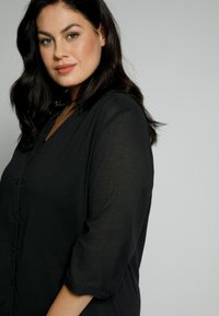 Schwarze Hemdbluse mit V-Ausschnitt, drei Viertel lange Ärmel und einem leichten, durchsichtigen Material. Einfaches Design ohne Muster.