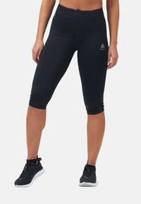 Schwarze Capri-Leggings mit hohem Bund, glatter Textur und kleinem Logo auf der Vorderseite. Getragen mit schwarzen Sportschuhen.