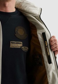 Teilweise sichtbare Person, die einen schwarzen PME Legend Pullover trägt und eine hellfarbige Jacke öffnet, in der ein PME Legend Etikett zu sehen ist.