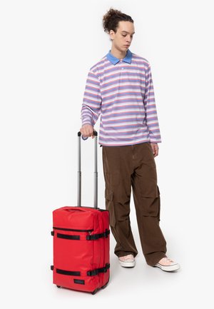 Roter Koffer mit schwarzen Riemen, rechteckig geformt und mit glatter Oberfläche. Modell trägt ein gestreiftes Langarmshirt und Cargo-Hosen.