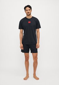 T-shirt de sport noir à manches courtes, avec un patch logo rouge sur la poitrine, associé à un short noir assorti. Tissu lisse, coupe décontractée.