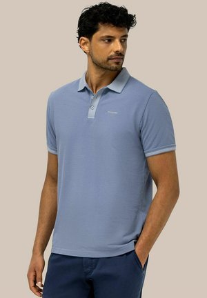 Man wearing light blue short-sleeve polo shirt with logo on chest and navy blue pants, standing with one hand in pocket, looking to side.