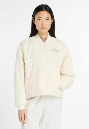 Lichtbeige bomberjack met een ritssluiting aan de voorkant, geribde manchetten en "New Balance Athletics" subtiel in het groen geborduurd op de borst.