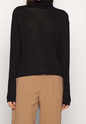 Personne portant un pull à col roulé noir à manches longues et un pantalon beige taille haute sur un fond clair uni.