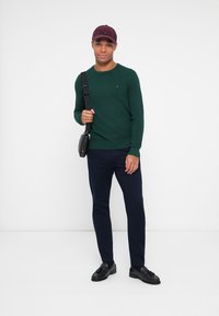 Maglione verde, pantaloni blu scuro e scarpe nere. L'individuo tiene una piccola borsa nera e indossa un cappellino bordeaux, con una texture di tessuto liscia.