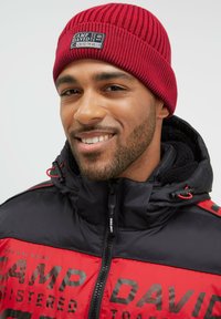 Red ribbed beanie with a branded patch, paired with a black and red puffer jacket featuring a zip closure and logo details.