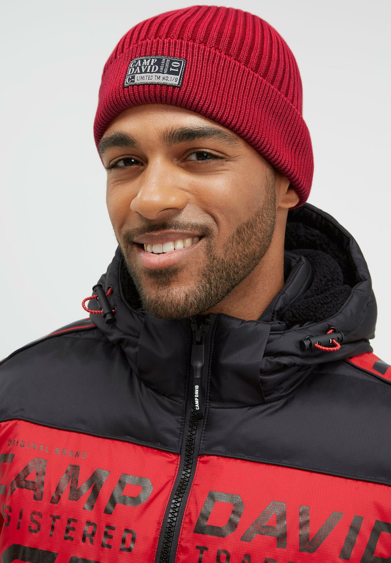 Red ribbed beanie with a branded patch, paired with a black and red puffer jacket featuring a zip closure and logo details.