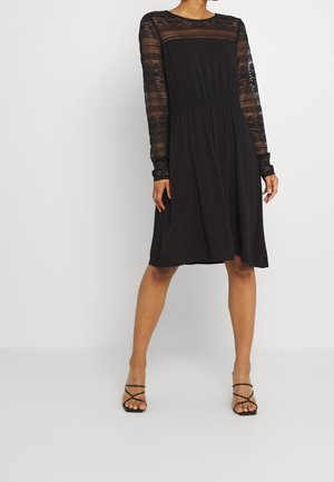 Kvinde iført en knælængde sort kjole med gennemsigtige blonder på lange ærmer og høj hals, kombineret med sorte remsandaler.