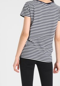Camiseta de algodón a rayas en azul marino y blanco, con mangas cortas y escote redondeado, combinada con pantalones ajustados negros. La vista trasera muestra un corte relajado.
