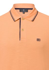 Jasnopomarańczowa koszulka polo z granatowymi akcentami. Posiada dwa guziki przy kołnierzyku oraz haftowane logo po lewej stronie na piersi. Teksturowana tkanina.