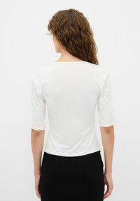 Top blanco ajustado con cuello redondo y mangas tres cuartos, fabricado con una tela suave, mostrado desde la parte posterior contra un fondo simple.