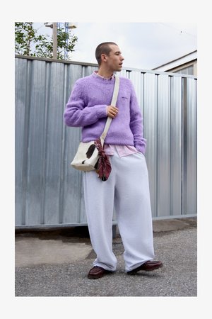 Lilla fuzzy sweater, lyserød skjorte under, grå løse sweatpants, mønstret taske med et maroon tørklæde, brune sko, står foran en metallisk baggrund.