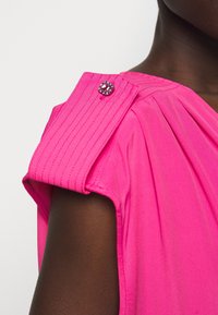 Haut sans manches rose vif avec un rabat d'épaule matelassé fixé par un petit bouton décoratif en gemme violet sur une peau foncée.