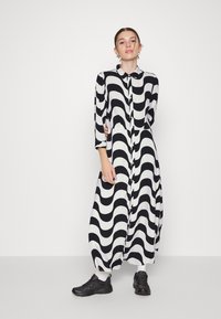 Vestido longo com padrão ondulado em preto e branco, com botão à frente, colarinho e mangas a três quartos, combinado com ténis pretos.