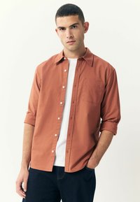 Chemise à boutons couleur rouille avec une poche, manches retroussées, portée sur un T-shirt blanc, associée à un jean en denim foncé.