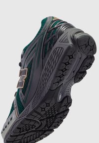 Groene en grijze hardloopschoen gekanteld zodat de zwarte, textuurrijke zool met hexagonaal profiel en het merklogo aan de zijkant zichtbaar zijn.