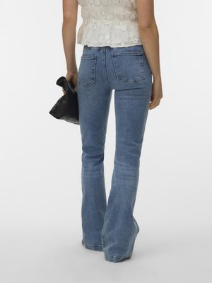 Személy világoskék bőszárú farmert és fehér ujjatlan felsőt visel, fekete clutch táskát tart a kezében, hátulnézetből látható.