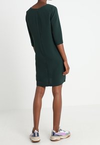 Dunkelgrünes, knielanges Kleid mit Dreiviertelärmeln und Reißverschluss auf der Rückseite. Kombiniert mit bunten Sportschuhen.