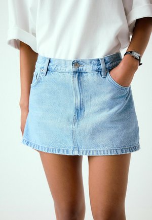 Femme portant une mini-jupe en jean bleu clair et un t-shirt blanc, avec une main dans la poche de la jupe et une montre noire au poignet gauche.