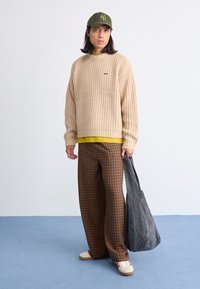 Suéter de malha bege, camisola amarela por baixo, calças xadrez castanhas e laranja, mala cinza, sapatos brancos com detalhes vermelhos, boné verde com logotipo.