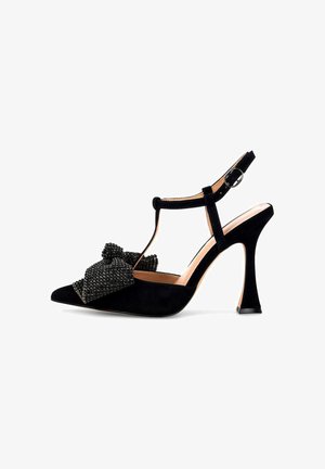 Zapato de tacón alto de ante negro con punta afilada, tira en forma de T y detalle de moño embellecido con cuentas negras en la puntera, que presenta un tacón esculpido.