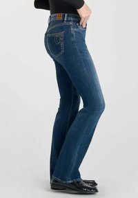 Jeans de campana azul oscuro con una cintura de media altura, bolsillos delanteros y traseros, y detalles cosidos. Parche de cuero de marca visible en la cinturilla.