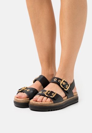 Pieds portant des sandales en cuir noir avec des boucles et œillets dorés, semelle en liège texturé et épais semelles noires nervurées.
