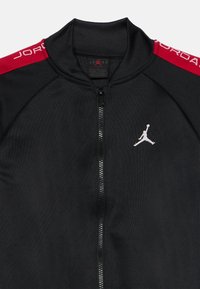 Chaqueta negra con cremallera, con franjas rojas en los hombros y el logo blanco de Jumpman en el pecho, con el texto "JORDAN" en los hombros.