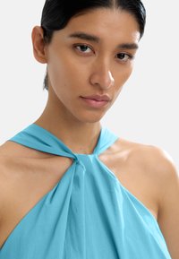 Jasnoniebieski top na ramiączkach z związanym przodem, wykonany z miękkiego materiału, o gładkiej teksturze i falbaniastym fasonie wokół dekoltu.