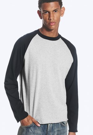 Giovane uomo che indossa una maglia stile baseball a maniche lunghe con maniche nere e corpo grigio chiaro, e jeans blu, in piedi con una mano in tasca.