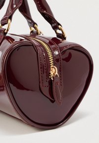 Sac à main en cuir verni bordeaux en forme de cœur, avec une fermeture éclair dorée, deux poignées et une finition polie, présentant des détails de couture subtils.