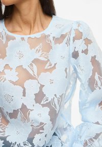Femme portant un chemisier bleu clair transparent avec une grande broderie florale en dentelle et de longues manches bouffantes, laissant apparaître la peau en dessous.