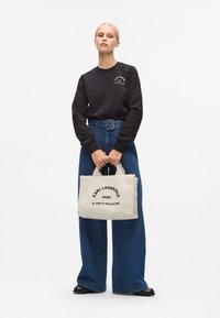 Zwarte sweatshirt, hoog getailleerde denim broek en crèmekleurige handtas van textuurstof met zwarte tekst: "KARL LAGERFELD PARIS."