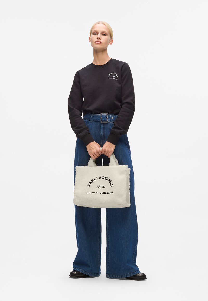 Zwarte sweatshirt, hoog getailleerde denim broek en crèmekleurige handtas van textuurstof met zwarte tekst: "KARL LAGERFELD PARIS."