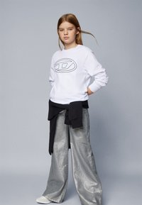Felpa bianca con logo nero, abbinata a pantaloni larghi argentati metallizzati. Maglione nero annodato in vita. Trame lisce.