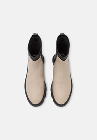 Botas de tobillo de cuero beige con un acento negro, que presentan una textura suave y lados elásticos, diseñadas para facilitar su uso y comodidad.