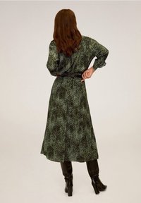 Abito lungo con stampa di serpente verde, con vita cinturata e maniche lunghe, caratterizzato da una silhouette fluida e una texture morbida. Stivali neri fino al ginocchio.
